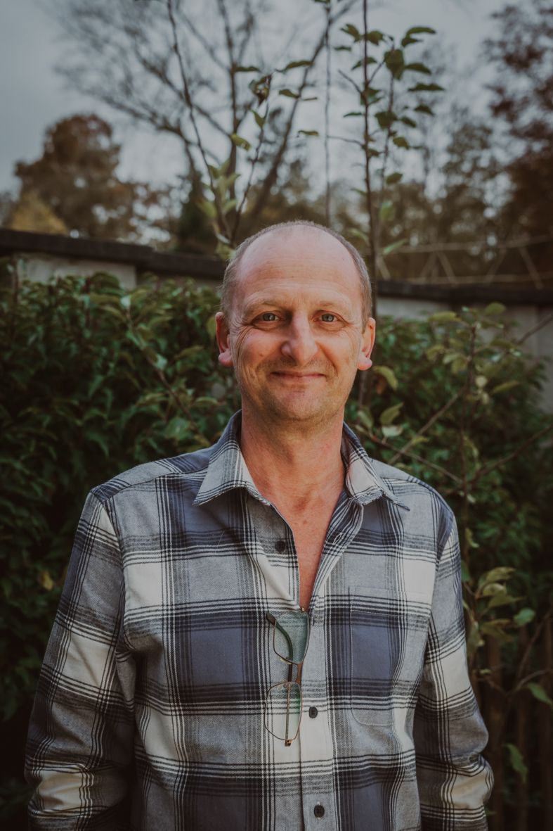 Persoon in geblokt overhemd lacht warm buiten bij groen gebladerte