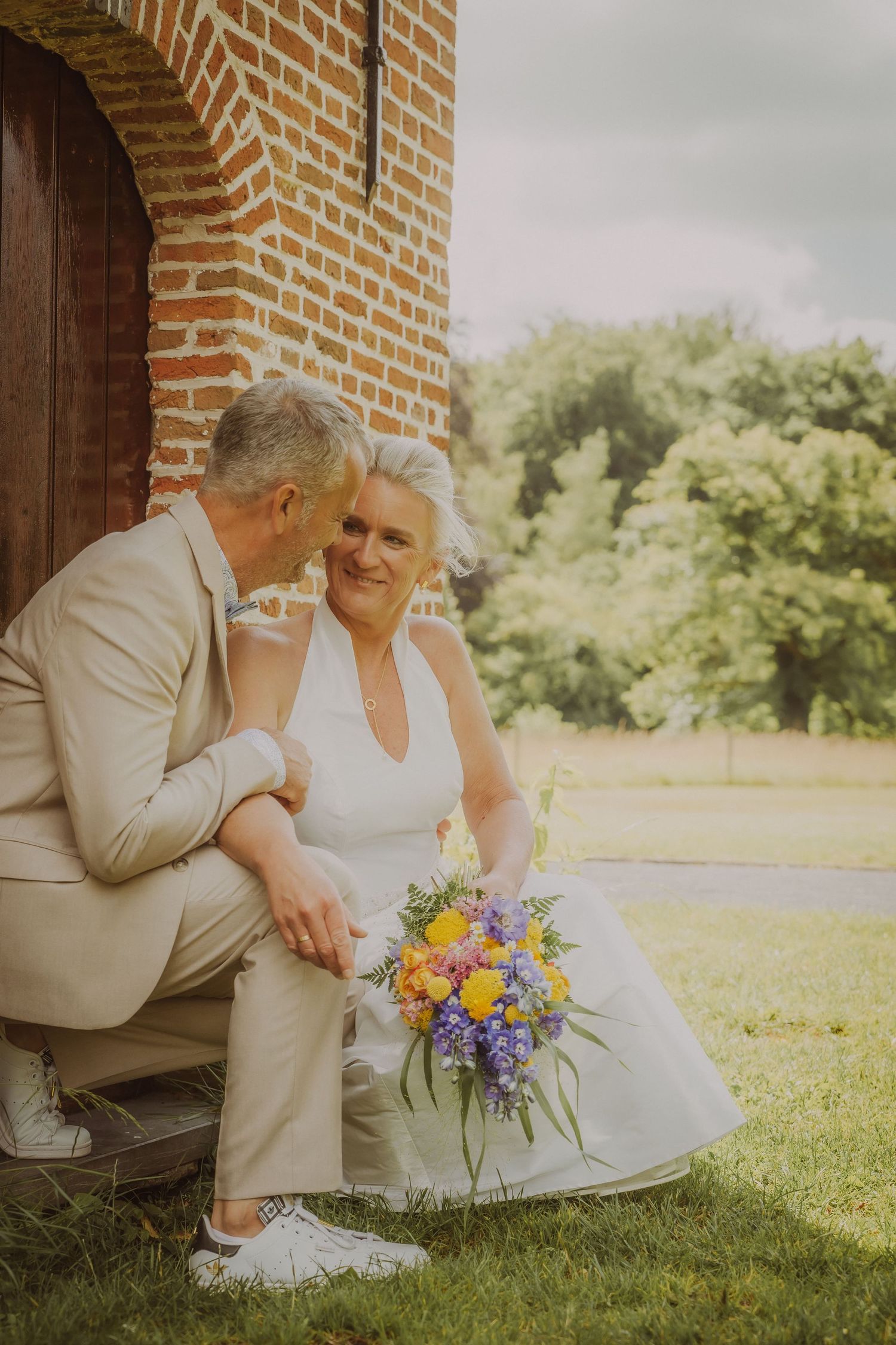 Koppel zit samen bij een bakstenen muur met kleurrijk bruiloftsboeket