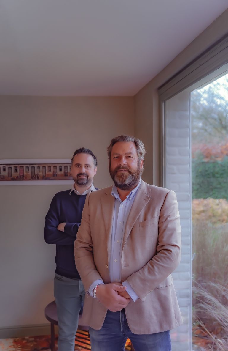 Twee mannen staan bij een raam, één in beige blazer, één in donkere kleding