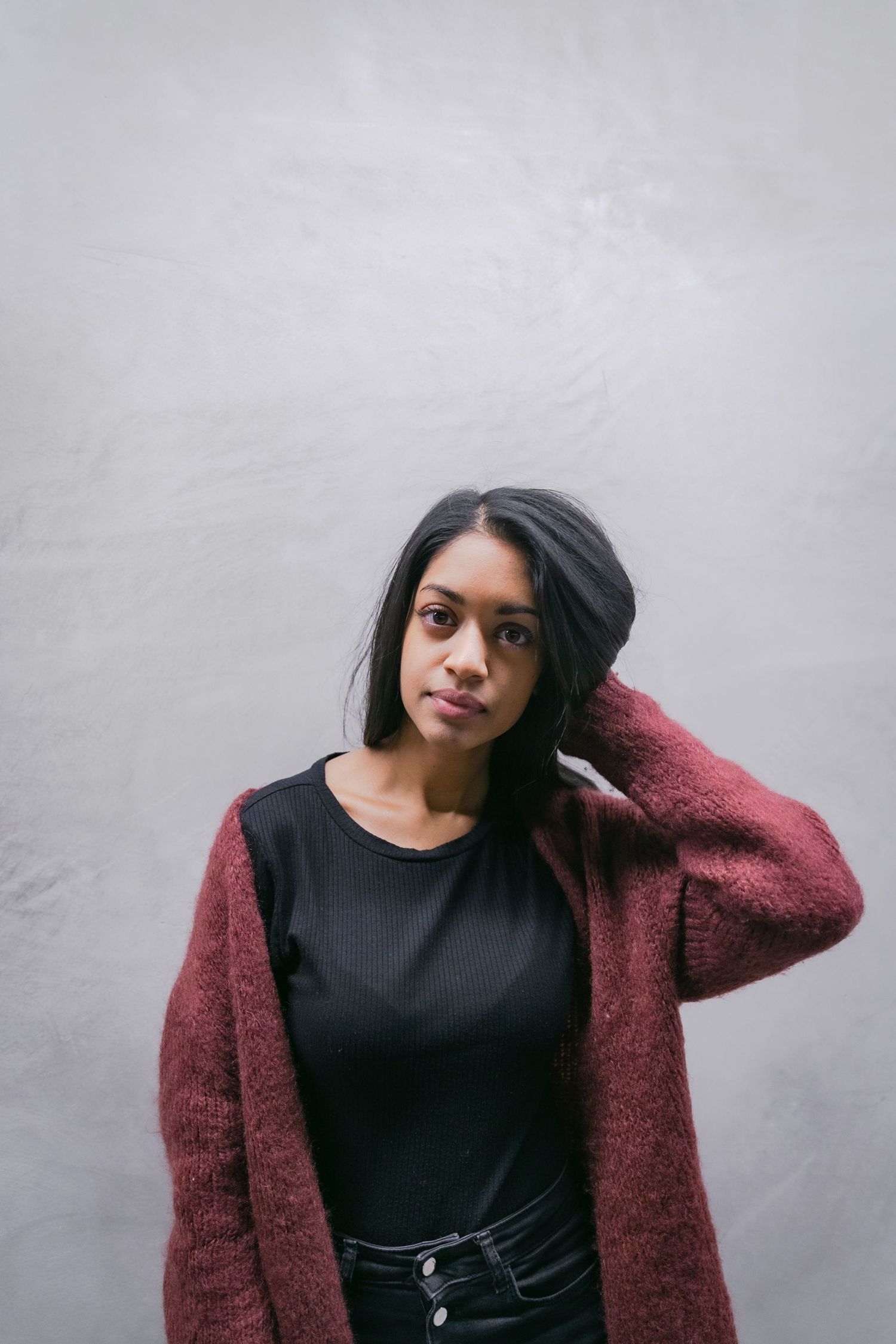 Vrouw in bordeaux vest en zwarte top poseert voor lichtgrijze muur